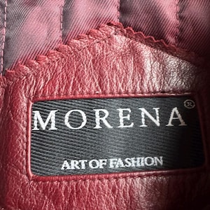 K&ouml;nnte beinhalten: Ein bordeauxrotes Lederkleidungsst&uuml;ck mit einem schwarzen, rechteckigen Stoffetikett mit dem wei&szlig;en Text MORENA und ART OF FASHION. Das Leder hat eine sichtbare Narbenstruktur und ist mit einem dunkelroten, vertikal gesteppten Innenfutter vern&auml;ht.