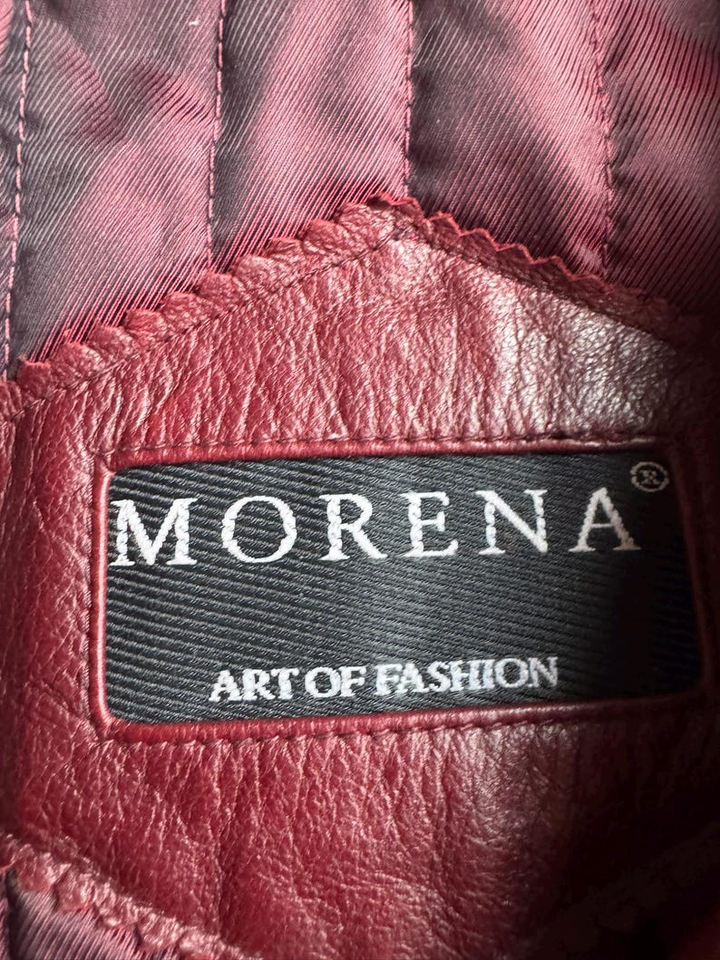 K&ouml;nnte beinhalten: Ein bordeauxrotes Lederkleidungsst&uuml;ck mit einem schwarzen, rechteckigen Stoffetikett mit dem wei&szlig;en Text MORENA und ART OF FASHION. Das Leder hat eine sichtbare Narbenstruktur und ist mit einem dunkelroten, vertikal gesteppten Innenfutter vern&auml;ht.