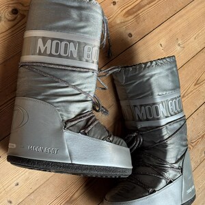 May include: A pair of silver Moon Boot winter boots. The boots have a shiny, metallic finish with the brand name printed in white across the top. They feature a lace-up design and a thick, black sole. The boots are designed for cold weather.