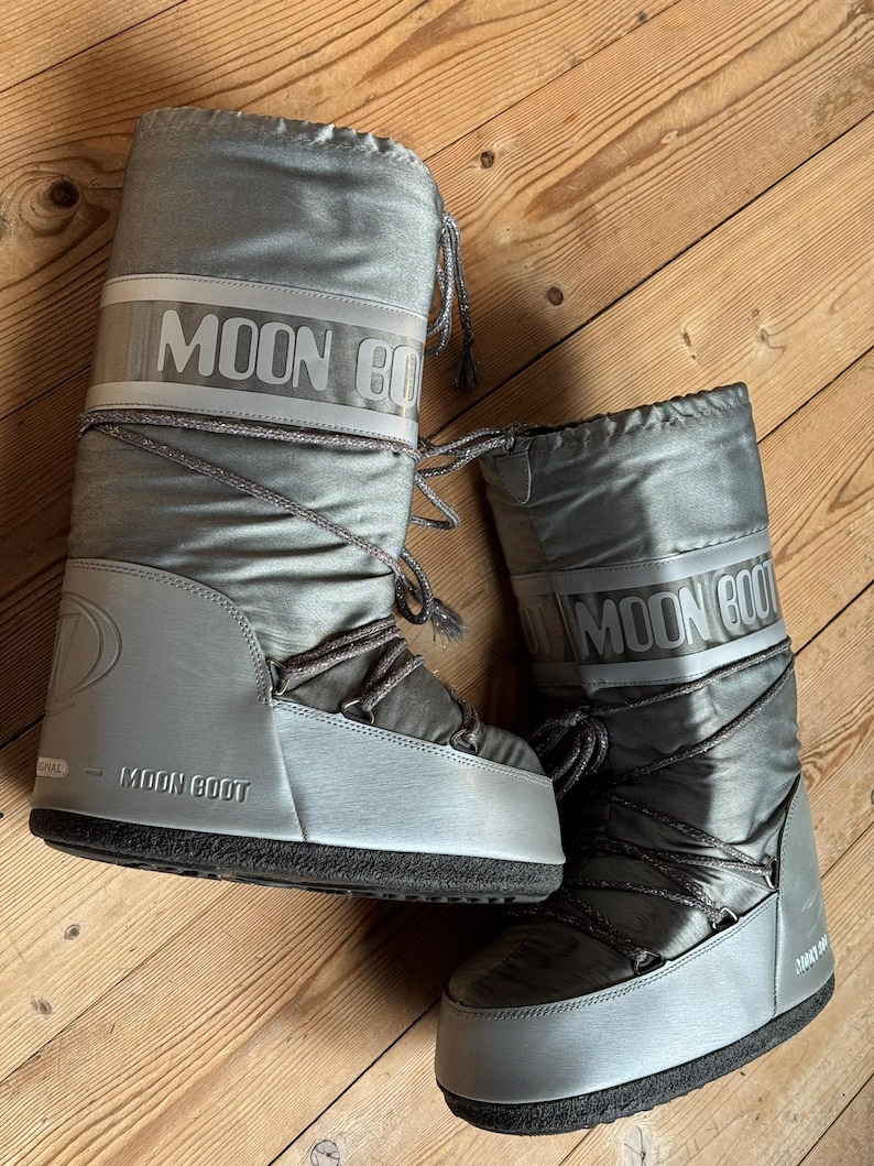 May include: A pair of silver Moon Boot winter boots. The boots have a shiny, metallic finish with the brand name printed in white across the top. They feature a lace-up design and a thick, black sole. The boots are designed for cold weather.