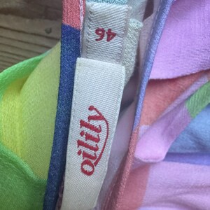 Pu&ograve; includere: Primo piano di un tessuto colorato con un'etichetta che recita "oilily" in corsivo rosso. Il tessuto &egrave; un patchwork di colori pastello tra cui rosa, blu, verde e giallo. L'etichetta riporta anche il numero "46".