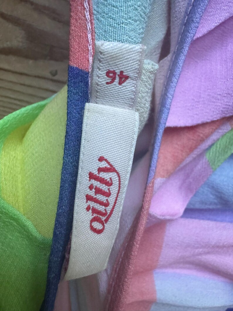 Pu&ograve; includere: Primo piano di un tessuto colorato con un'etichetta che recita "oilily" in corsivo rosso. Il tessuto &egrave; un patchwork di colori pastello tra cui rosa, blu, verde e giallo. L'etichetta riporta anche il numero "46".