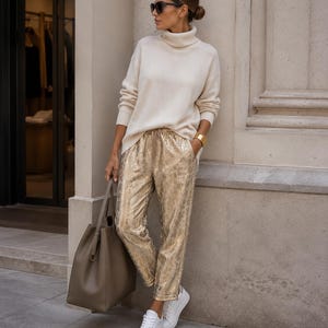 Puede incluir: Un suéter de cuello alto color crema combinado con pantalones dorados metalizados con un sutil patrón texturizado. El conjunto incluye zapatillas blancas de cuero, un bolso grande color oliva, gafas de sol oscuras y un brazalete dorado ancho.