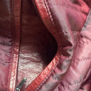 K&ouml;nnte beinhalten: Das Innere einer tiefroten Ledertasche mit gestepptem Stofffutter in einem passenden dunklen Burgunderrot. An der Seite befindet sich eine Rei&szlig;verschlusstasche mit einem silberfarbenen Metallzipper. Ein kleines schwarzes Etikett ist sichtbar.