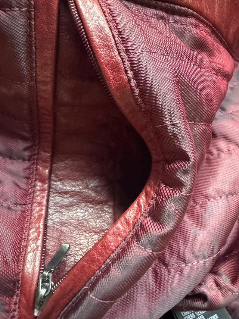 K&ouml;nnte beinhalten: Das Innere einer tiefroten Ledertasche mit gestepptem Stofffutter in einem passenden dunklen Burgunderrot. An der Seite befindet sich eine Rei&szlig;verschlusstasche mit einem silberfarbenen Metallzipper. Ein kleines schwarzes Etikett ist sichtbar.
