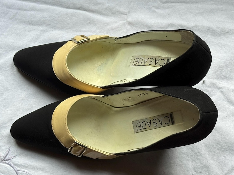 May include: A pair of black Casadei pumps with a pointed toe and a beige strap across the top, secured with a small buckle. The brand name is visible inside the shoe.