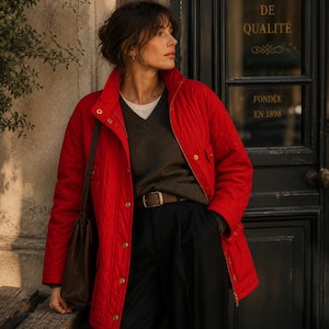 May include: A quilted red jacket with a high collar and gold-colored buttons. The jacket is worn over a dark green sweater and black pants. A brown leather bag hangs from the shoulder. The image is taken outdoors.