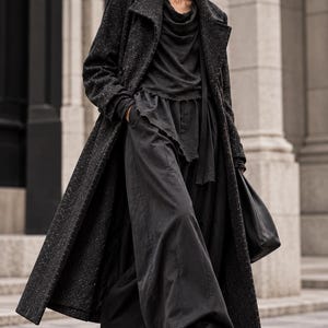 May include: A long, dark gray speckled wool-blend coat worn over a black cowl-neck top with raw-edge detailing and matching wide-leg trousers. The outfit includes black leather shoes and a black leather handbag, paired with dark sunglasses.