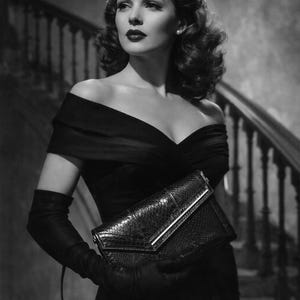 Op de afbeelding: Zwart-witfoto van een vrouw in een zwarte off-the-shoulder jurk, lange handschoenen en een clutch met textuur. De vrouw heeft donker, golvend haar en staat bij een trap.