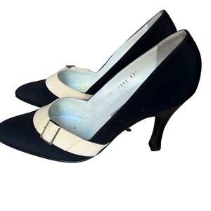 May include: A pair of black pumps with a cream-colored strap and buckle detail. The heels are slender and dark, and the shoes have a pointed toe. The interior of the shoe is white.