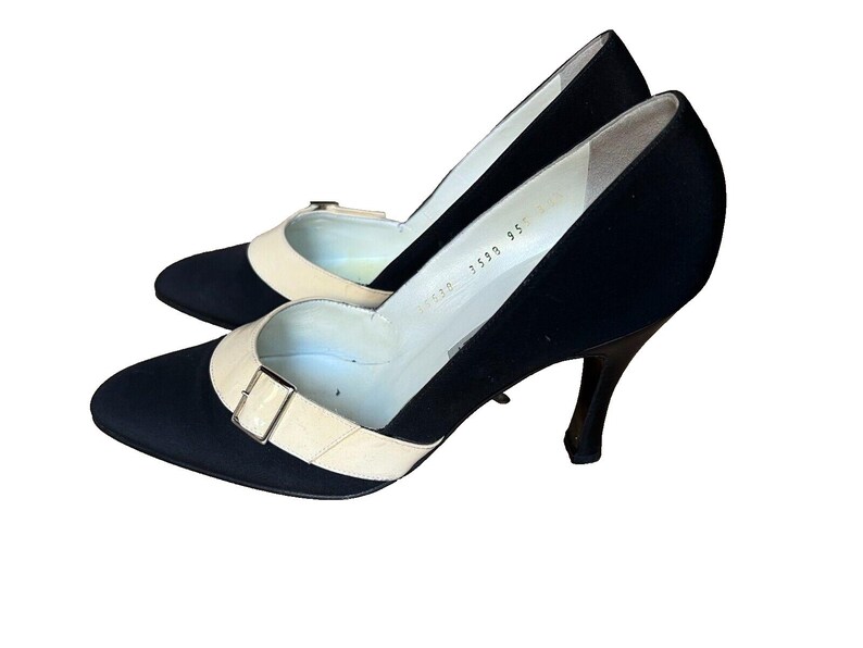 May include: A pair of black pumps with a cream-colored strap and buckle detail. The heels are slender and dark, and the shoes have a pointed toe. The interior of the shoe is white.