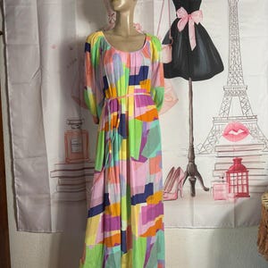Pu&ograve; includere: Un abito lungo maxi colorato a maniche lunghe con un motivo geometrico nei toni del rosa, verde, blu e arancione. L'abito ha uno scollo tondo e una cintura abbinata. L'abito &egrave; esposto su un manichino.