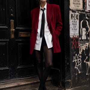Könnte beinhalten: Ein dunkelroter Blazer über einem weißen Hemd mit Knopfleiste und einer schmalen schwarzen Krawatte. Das Outfit umfasst eine transparente schwarze Strumpfhose und schwarze Slipper. Der Blazer hat betonte Schultern und eine zweireihige Knopfleiste.