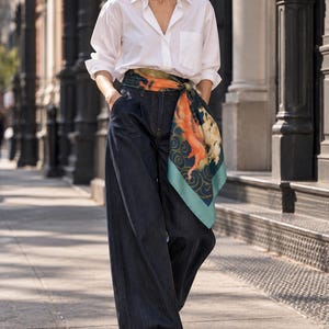 Puede incluir: Una camisa blanca de botones con bolsillo en el pecho, combinada con pantalones vaqueros de pierna ancha en color azul índigo oscuro. Un pañuelo de seda cuadrado con un patrón gráfico en naranja, verde azulado y crema atado a la cintura.