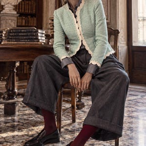 May include: A light green textured jacket with a scalloped cream trim, a grey collared shirt, wide-leg charcoal grey trousers, dark red ribbed socks, and black leather loafers. The outfit is accessorised with dark-tinted sunglasses.
