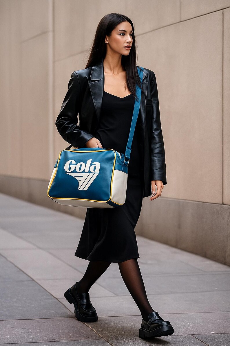 May include: A person wearing a black dress, black leather jacket, and black shoes carries a blue and white Gola shoulder bag. The bag has a white and yellow trim and the Gola logo.