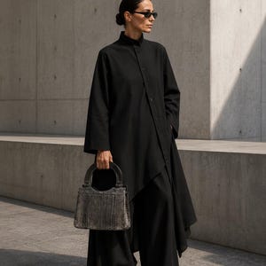 Può includere: Una camicia nera a maniche lunghe con bottoni e orlo asimmetrico, abbinata a pantaloni neri a gamba larga e scarpe nere a punta. Una borsa strutturata grigia con motivo a pelle di serpente e manico superiore arrotondato è tenuta in mano.