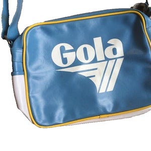 May include: A blue shoulder bag with the word "Gola" in white block letters. The bag has a white and yellow trim, and a blue shoulder strap. The bag is made of a synthetic material.