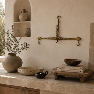 Op de afbeelding: Een interieurscène in neutrale tinten met diverse decoratieve items. Een messing wandweegschaal hangt boven een stenen aanrecht. Een vaas met groen, kommen, een kaars en boeken zijn op het aanrecht geplaatst.