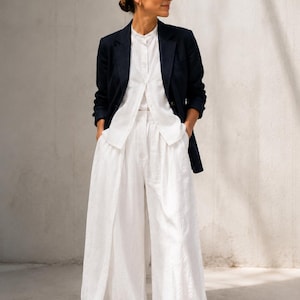 Puede incluir: Un blazer azul marino oscuro sobre una camisa blanca abotonada y pantalones blancos de pierna ancha. El conjunto incluye gafas de sol redondas y zapatos blancos. El atuendo presenta una chaqueta estructurada con pantalones holgados.