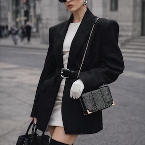 Könnte beinhalten: Eine Frau in einem schwarzen Blazer, einem weißen Kleid und schwarzen Stiefeln. Sie trägt Sonnenbrille, Ohrringe und weiße Handschuhe. Eine schwarze Handtasche mit goldener Kette und eine schwarze Tragetasche mit "BALENCIAGA" sind sichtbar.