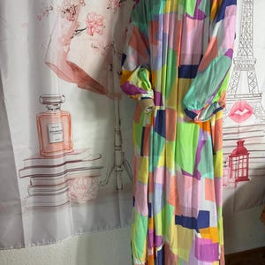 Pu&ograve; includere: Un abito lungo e fluido con un colorato motivo geometrico. L'abito presenta un mix di colori vivaci tra cui rosa, verde, giallo e blu. Ha maniche lunghe e scollatura rotonda. L'abito sembra essere realizzato in un tessuto leggero.