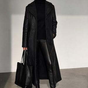 Könnte beinhalten: Ein langer schwarzer Ledermantel mit breitem Kragen, getragen über einem schwarzen Rollkragenpullover und einer schwarzen Lederhose. Das Outfit umfasst schwarze Lederstiefeletten mit dicker Sohle und eine große schwarze Ledertragetasche.