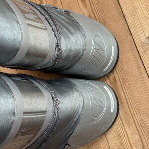 May include: A pair of silver-gray snow boots with the brand name "MN" embossed on the toe and upper shaft. The boots have a shiny, metallic finish and a drawstring closure. The soles are black.