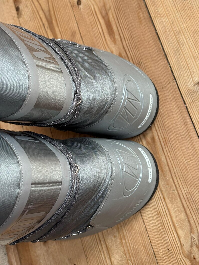 May include: A pair of silver-gray snow boots with the brand name "MN" embossed on the toe and upper shaft. The boots have a shiny, metallic finish and a drawstring closure. The soles are black.