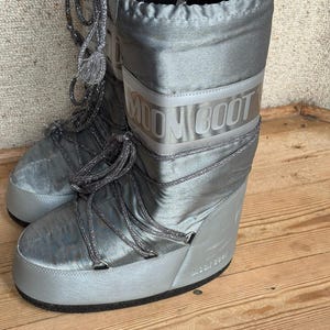 May include: A pair of silver Moon Boots with the brand name printed on the side. The boots have a shiny, metallic finish, with silver laces and a thick, rounded sole. The boots are designed for cold weather.