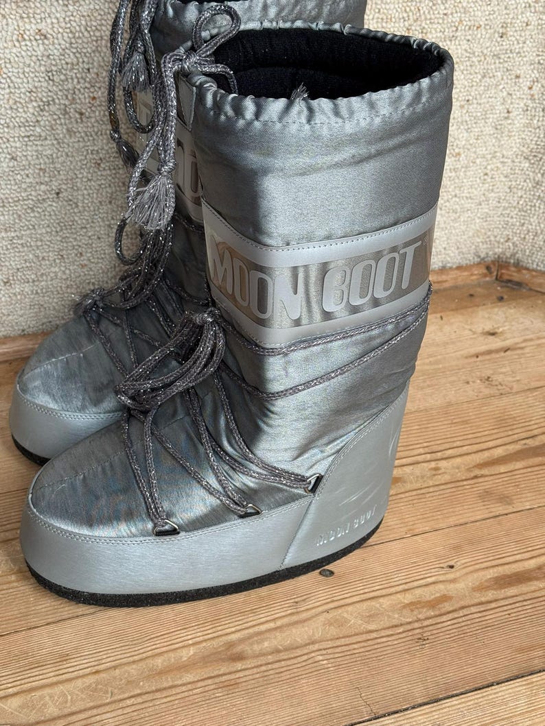 May include: A pair of silver Moon Boots with the brand name printed on the side. The boots have a shiny, metallic finish, with silver laces and a thick, rounded sole. The boots are designed for cold weather.