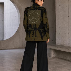 Op de afbeelding: Een olijfgroene blazer met een zwart geometrisch patroon en een groot vierkant embleem op de rug. Het embleem toont een wit gestippeld vredesteken. De blazer wordt gecombineerd met een zwarte wijde broek en zwarte plateau-laarzen.