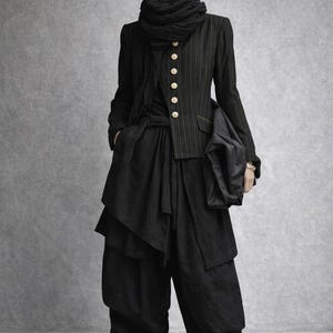 Könnte beinhalten: Eine Schaufensterpuppe in einem schwarzen Ensemble. Das Outfit umfasst eine Baskenmütze, einen Schal, eine Nadelstreifenjacke mit goldenen Knöpfen, eine mehrlagige Hose und Stiefel. Die Jacke hat dünne goldene Streifen. Der Stil ist avantgardistisch und anspruchsvoll.