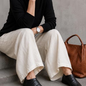 May include: A black long-sleeved sweater, wide-leg off-white trousers, and black leather slip-on shoes. A brown leather tote bag sits nearby. The ensemble includes a silver-toned wristwatch with a white face and a round metal link band.