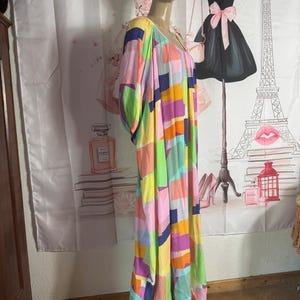 Pu&ograve; includere: Un abito lungo in stile patchwork dai colori vivaci, con scollo a V e maniche corte. L'abito presenta una vivace miscela di motivi rettangolari rosa, gialli, verdi, blu e arancioni. L'abito &egrave; esposto su un manichino.