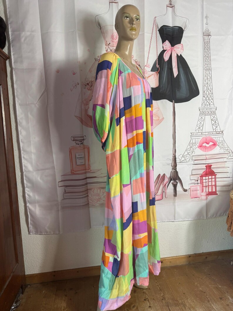 Pu&ograve; includere: Un abito lungo in stile patchwork dai colori vivaci, con scollo a V e maniche corte. L'abito presenta una vivace miscela di motivi rettangolari rosa, gialli, verdi, blu e arancioni. L'abito &egrave; esposto su un manichino.