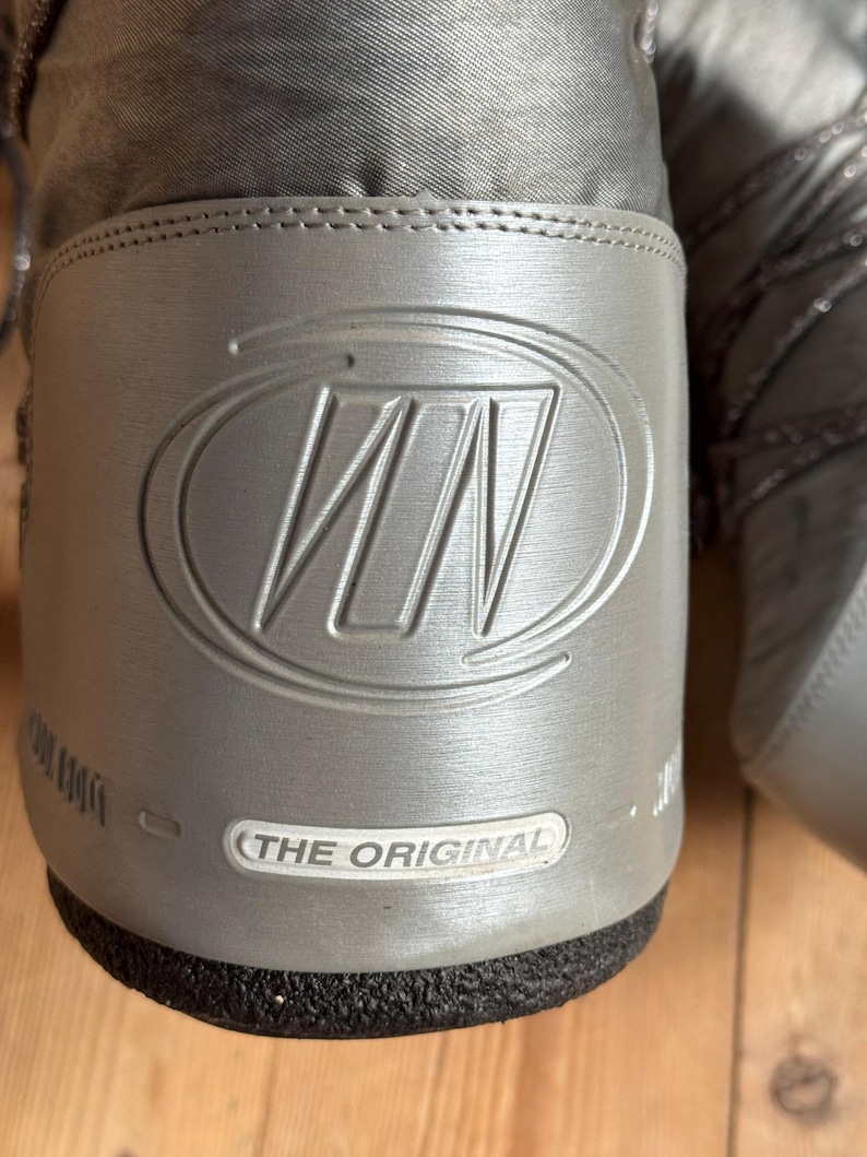 May include: Close-up of a gray winter boot with a silver logo and the text "THE ORIGINAL". The boot has a textured upper and a black sole. The logo is a stylized "WN" within a circle.