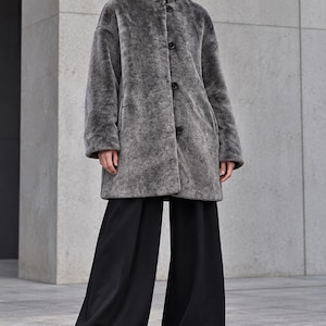 Könnte beinhalten: Ein grauer Kunstpelzmantel mit hohem Kragen und Knopfleiste vorne, kombiniert mit einer weiten schwarzen Hose und schwarzen Schuhen mit Profilsohle. Der Mantel hat eine strukturierte, flauschige Oberfläche und lange Ärmel für ein klares Design.