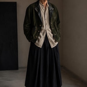 May include: An olive green suede jacket with black leather lapels and metallic buttons, worn over a beige button-down shirt and long black pleated trousers. The outfit is completed with black leather shoes, creating a layered ensemble.