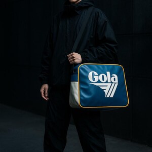 May include: A person wearing black sunglasses and a black hooded jacket, holding a blue, white, and yellow Gola shoulder bag. The bag features the Gola logo in white. The person is also wearing black pants and sneakers.