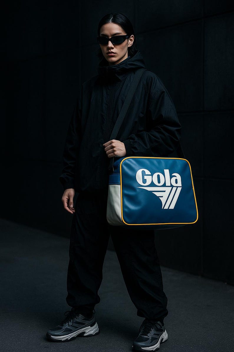 May include: A person wearing black sunglasses and a black hooded jacket, holding a blue, white, and yellow Gola shoulder bag. The bag features the Gola logo in white. The person is also wearing black pants and sneakers.