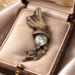Könnte beinhalten: Antik anmutende Brosche aus goldfarbenem Metall, verziert mit klaren Kristallen und einem großen, ovalen Mittelstein. Die Brosche liegt in einer braunen Vintage-Schmuckschatulle mit beigem Innenraum.