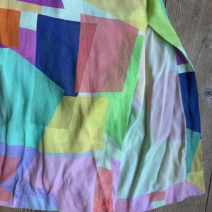 Pu&ograve; includere: Un capo colorato con un motivo geometrico. Il tessuto presenta rettangoli sovrapposti in tonalit&agrave; di viola, arancione, blu, verde, giallo e rosa. Il capo ha uno spacco laterale, che rivela un tessuto pi&ugrave; chiaro sottostante.