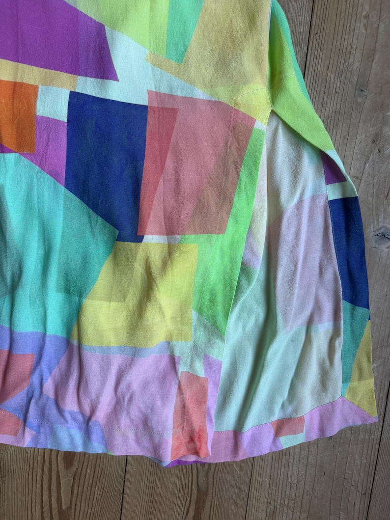 Pu&ograve; includere: Un capo colorato con un motivo geometrico. Il tessuto presenta rettangoli sovrapposti in tonalit&agrave; di viola, arancione, blu, verde, giallo e rosa. Il capo ha uno spacco laterale, che rivela un tessuto pi&ugrave; chiaro sottostante.