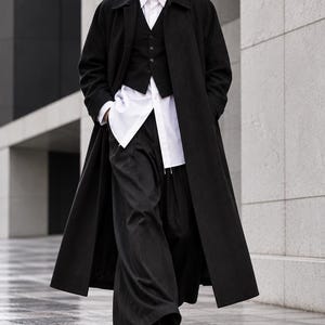 Puede incluir: Un abrigo largo negro sobre un chaleco negro, una camisa blanca de cuello rígido y pantalones negros de pernera ancha. El conjunto se completa con zapatos de cuero negro y gafas de sol oscuras, creando un estilo monocromático y elegante.