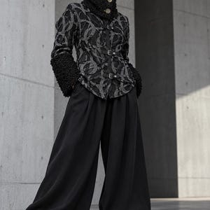 May include: A black and grey patterned jacket with a high collar, textured cuffs, and three front buckle closures, paired with wide-leg black trousers and black platform boots. The jacket features a repeating circular design in grey on black.