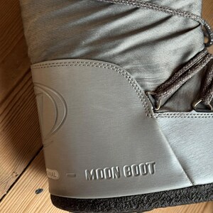May include: A close-up of a silver Moon Boot. The boot features a padded nylon upper with silver laces and a silver rubber sole. The brand name "MOON BOOT" is embossed on the side.