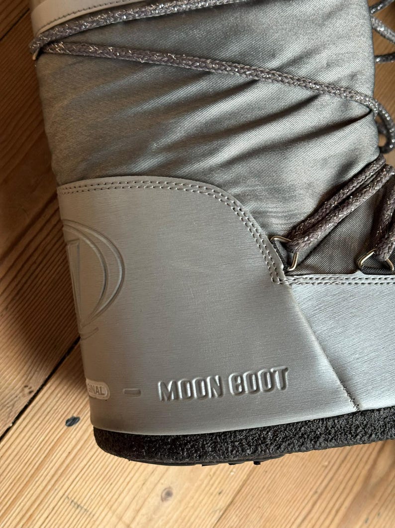 May include: A close-up of a silver Moon Boot. The boot features a padded nylon upper with silver laces and a silver rubber sole. The brand name "MOON BOOT" is embossed on the side.