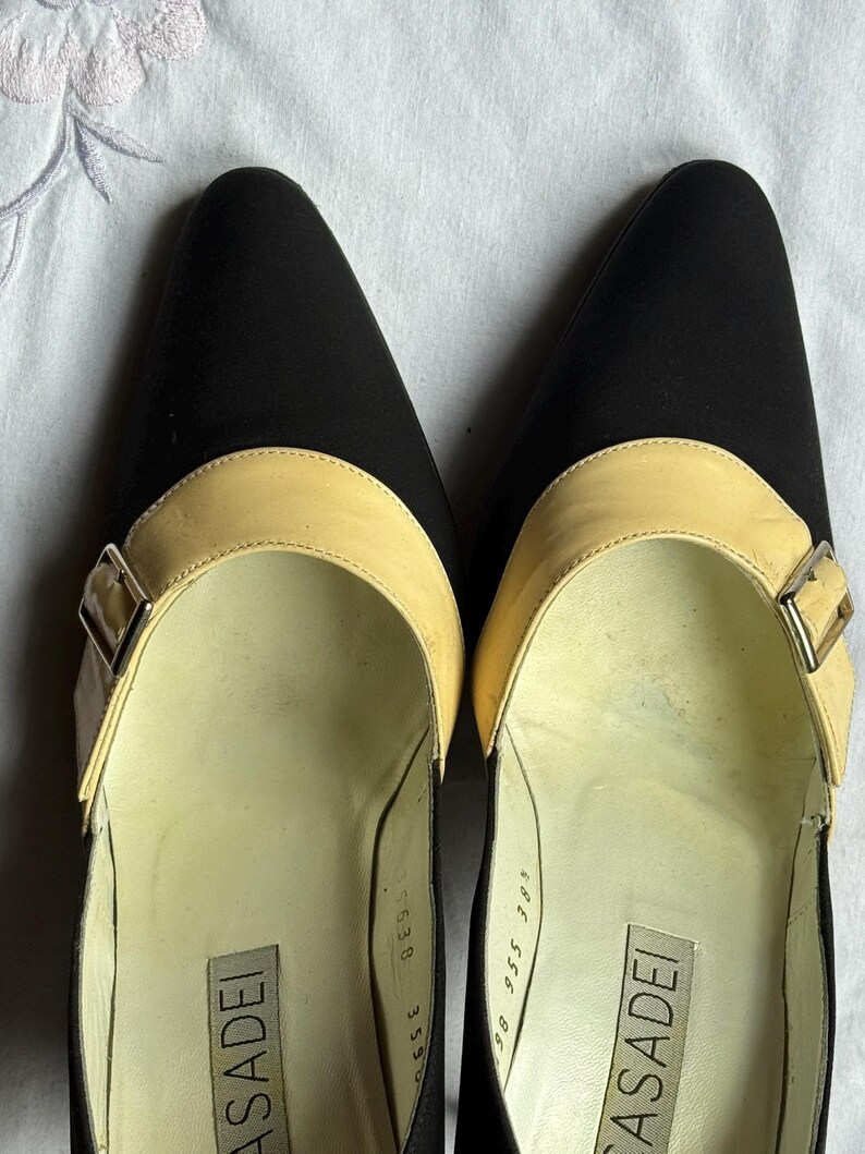 May include: Pair of black pointed-toe pumps with a light tan strap across the top, secured by a silver buckle. The brand name "CASADEI" is visible inside the shoe. The shoes have a classic, elegant design.