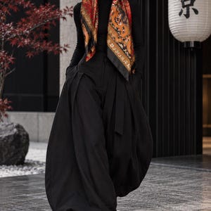 Puede incluir: Una camiseta negra de cuello alto y manga larga combinada con pantalones negros plisados de corte voluminoso y botas negras. Un pañuelo de seda rojo y naranja con motivos dorados se lleva sobre la cabeza, junto a gafas de sol negras.